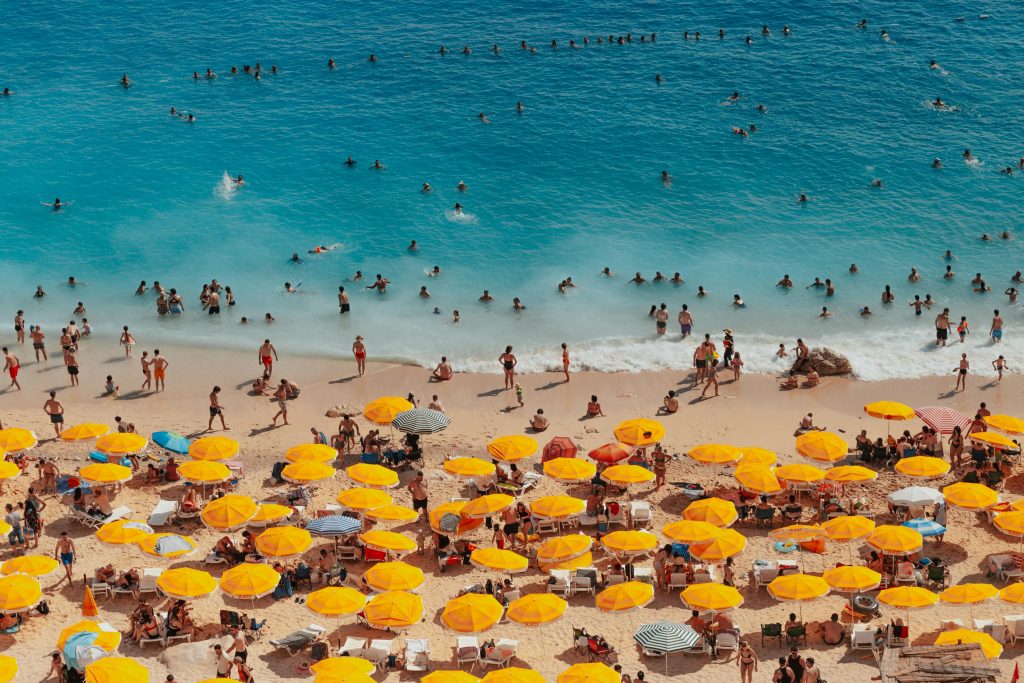 A vibrant beach scene in Kaş, Türkiye, with people enjoying the sea under yellow umbrellas.